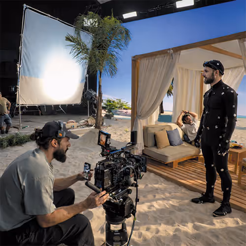 Film crew shooting a scene on a sandy set with a man in a motion capture suit and another man operating a camera.
