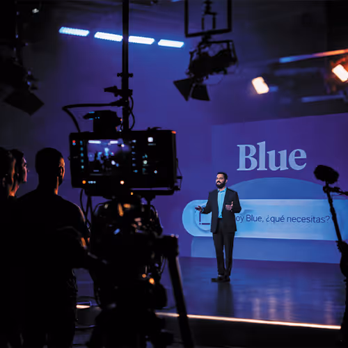 Man giving a presentation on stage with the word 'Blue' projected behind him, while camera crew films in a dark studio.