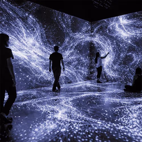 People interacting inside a dark room with immersive digital projections of swirling star-like lights on the walls and floor.
