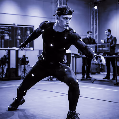 Man in a motion capture suit striking a pose in a studio with two people working in the background.